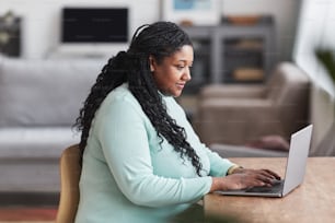 Seitenansichtsporträt einer kurvigen Afroamerikanerin, die einen Laptop am Schreibtisch benutzt und lächelt, während sie die Arbeit von zu Hause aus in minimalem Innenraum und Kopierraum genießt