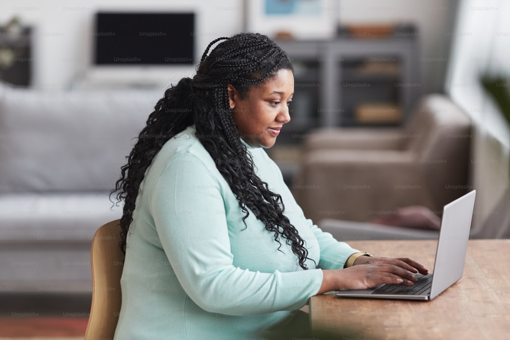 Retrato de vista lateral de una mujer afroamericana con curvas que usa una computadora portátil en el escritorio y sonríe mientras disfruta del trabajo desde casa en un interior mínimo, espacio de copia