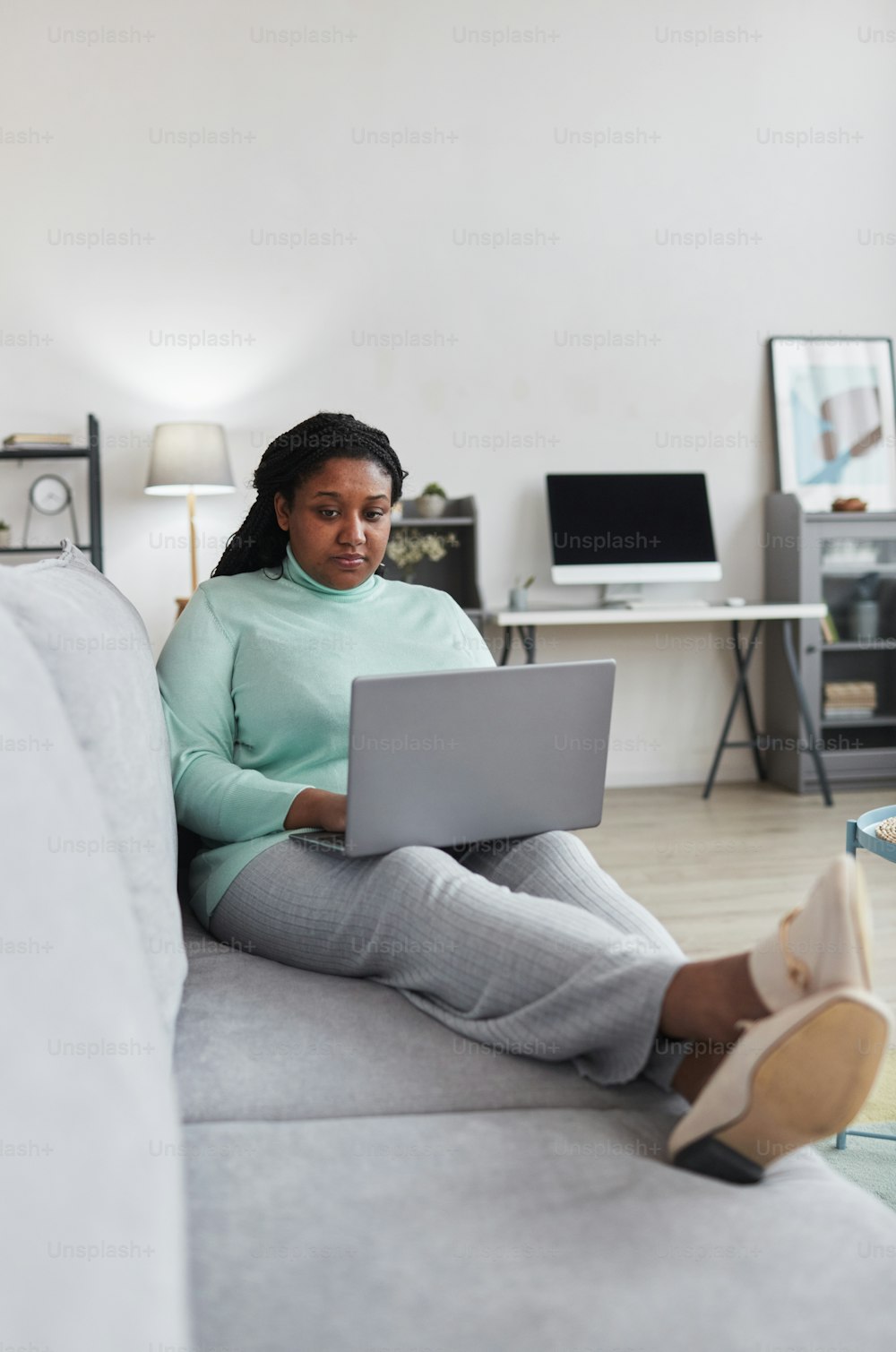 Vertikales Ganzkörperporträt einer kurvigen Afroamerikanerin, die einen Laptop benutzt, während sie die Arbeit von zu Hause aus genießt und sich auf der Couch in einem modernen, minimalistischen Interieur entspannt