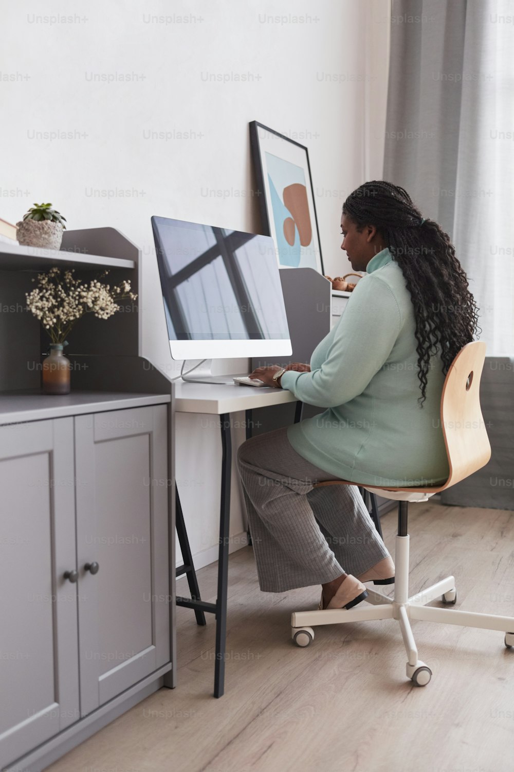 Vertikales Ganzkörperporträt einer kurvigen Afroamerikanerin, die den PC am Schreibtisch benutzt und die Arbeit von zu Hause aus in einem minimalistischen Innenraum genießt