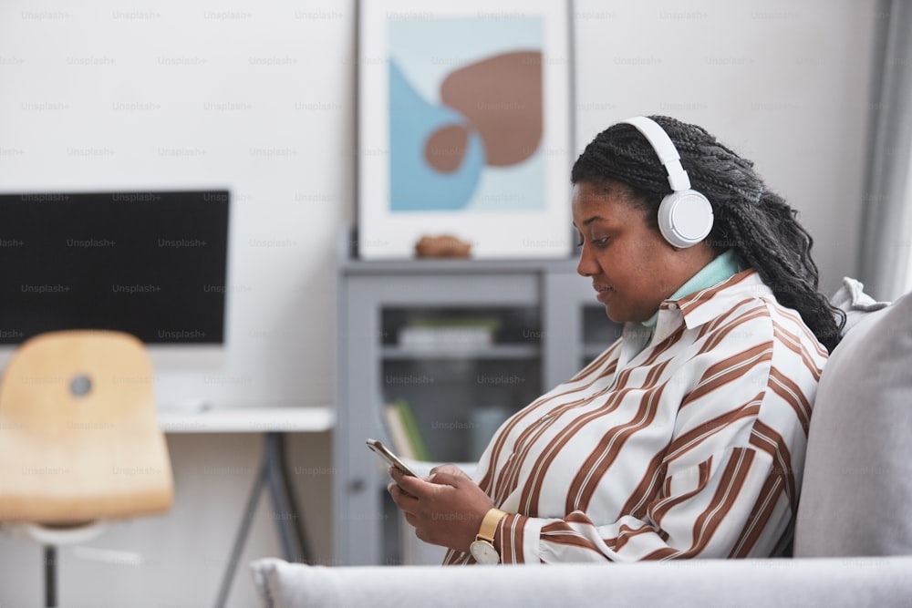Side view portrait of curvy African American woman wearing headphones and listening to music via smartphone while sitting on couch in minimal home interior, copy space