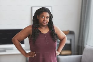 Retrato de cintura para arriba de una mujer afroamericana con curvas mirando a la cámara mientras está de pie con las manos en las caderas en el interior mínimo de la casa, copie el espacio