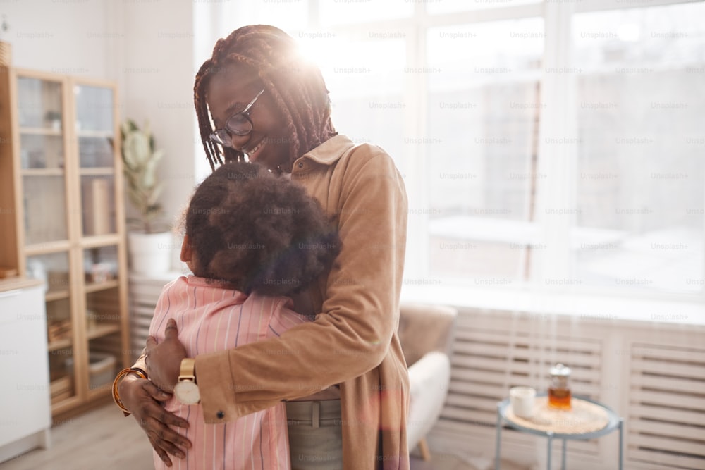 Waist up portrait of contemporary African-American mother embracing daughter while standing in cozy home interior lit by sunlight, copy space