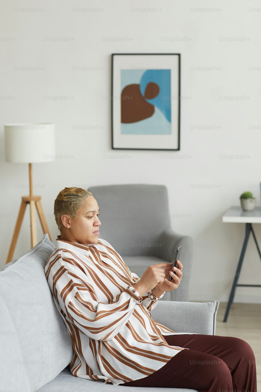 Vertikales Seitenansichtsporträt einer modernen gemischtrassigen Frau, die ein Smartphone benutzt, während sie auf dem Sofa in einem minimalen Wohnraum sitzt