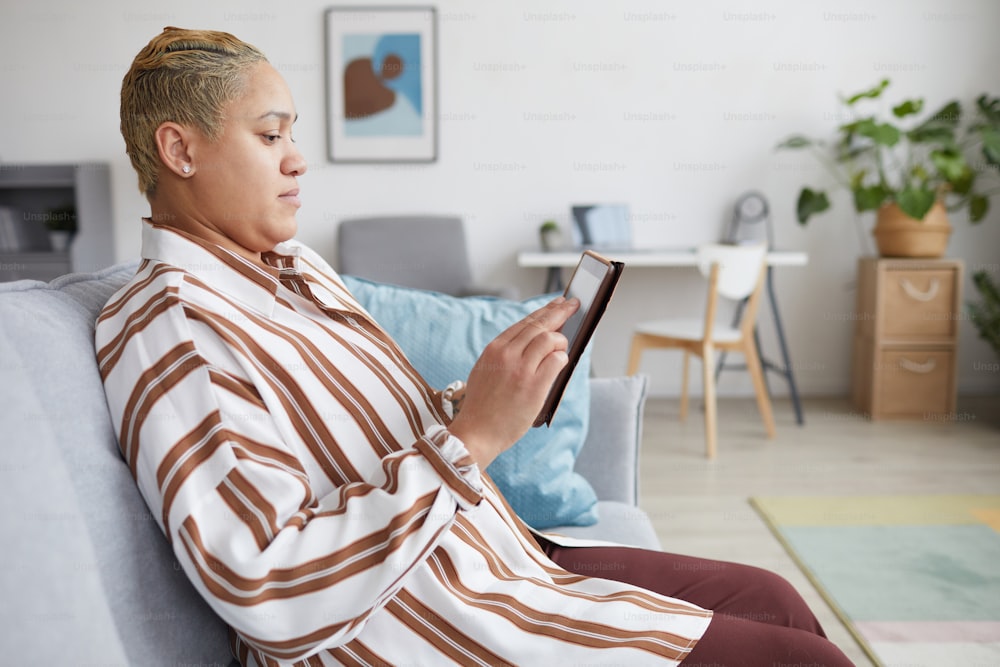 Vue latérale d’une femme métisse moderne tenant une tablette numérique et utilisant des services en ligne assise sur un canapé à la maison, espace de copie