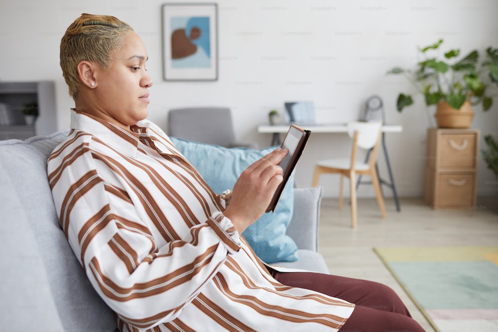 Seitenansichtsporträt einer modernen gemischtrassigen Frau, die ein digitales Tablet hält und Online-Dienste nutzt, während sie zu Hause auf dem Sofa sitzt, Kopierraum