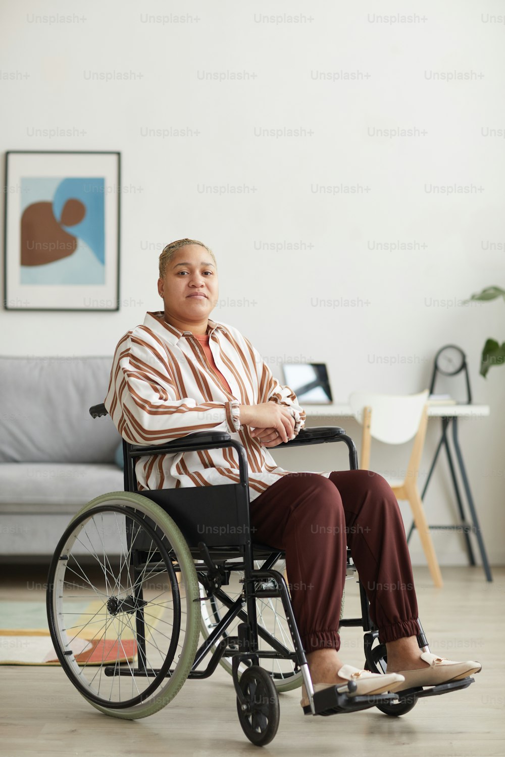 Vertikales Ganzkörperporträt einer modernen gemischtrassigen Frau, die im Rollstuhl sitzt und in minimalem Wohnraum in die Kamera schaut