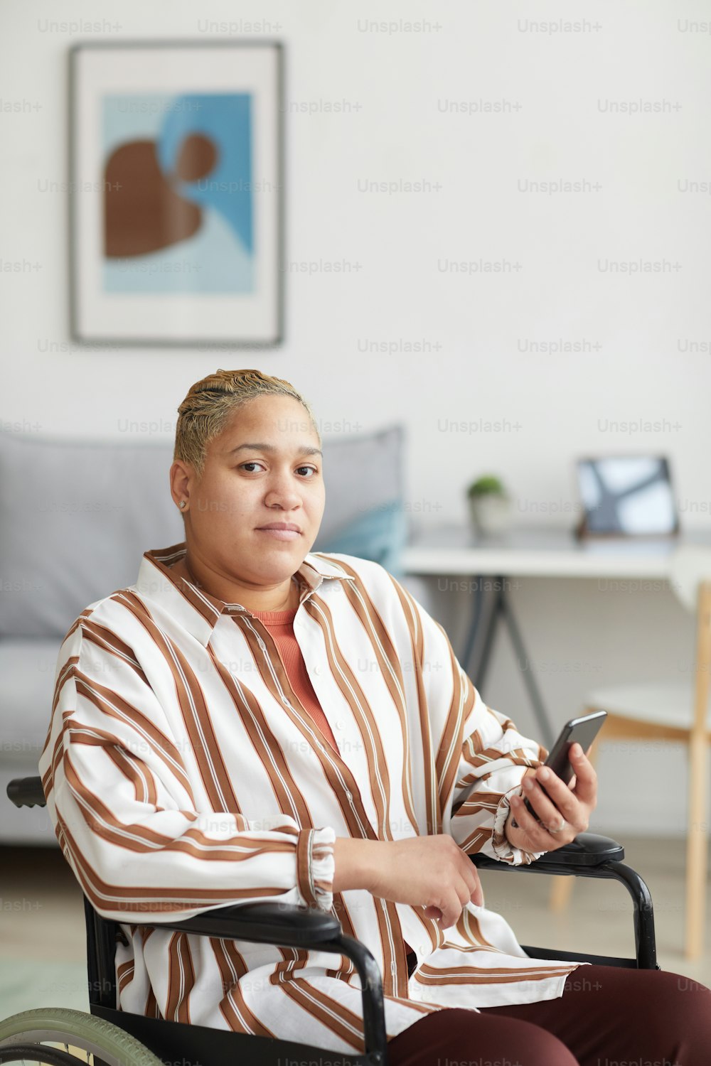 Portrait vertical d’une femme métisse moderne assise dans un fauteuil roulant et utilisant un smartphone tout en regardant l’appareil photo dans un intérieur minimal, espace de copie