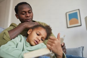 Portrait en contre-plongée d’un père et d’une fille afro-américains aimants lisant assis ensemble sur un canapé dans un intérieur confortable, espace de copie