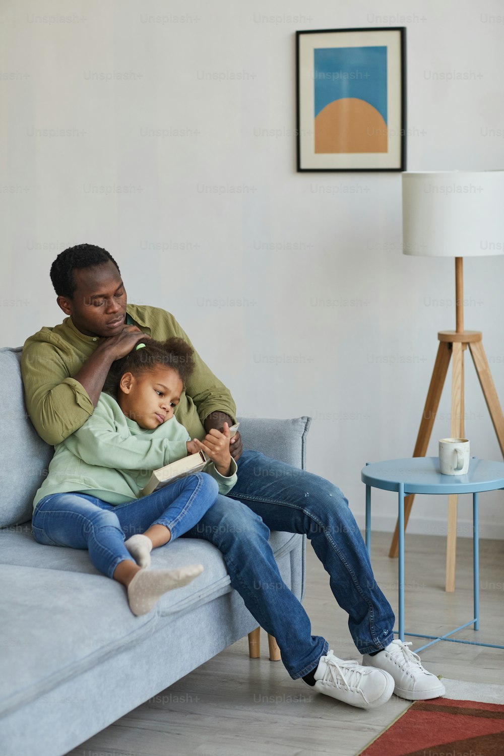 Ganzkörperporträt eines liebevollen afroamerikanischen Vaters und einer Tochter, die lesen, während sie zusammen auf der Couch in einem gemütlichen Wohnraum sitzen