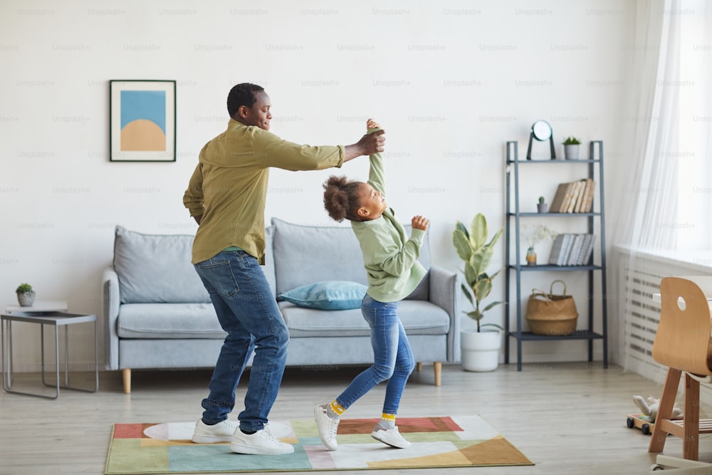 Retrato de cuerpo entero de un padre afroamericano feliz bailando con su hija mientras se divierte en el interior mínimo de la casa, espacio de copia