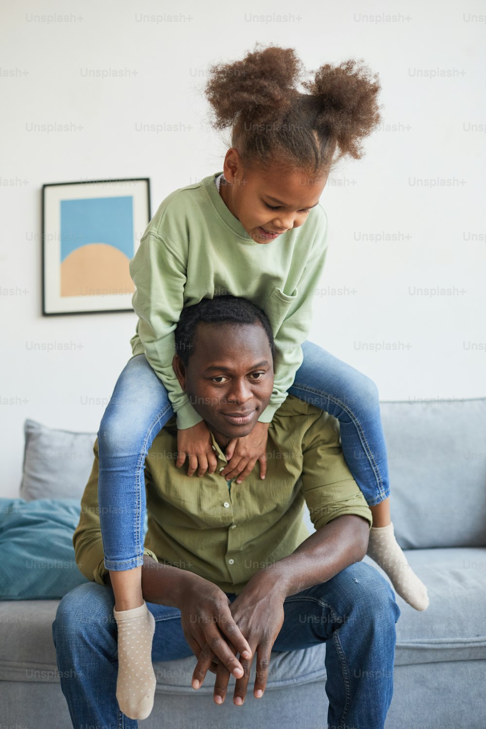 Vertikales Porträt eines niedlichen afroamerikanischen Mädchens, das auf den Schultern des Vaters sitzt, während es zusammen in einem gemütlichen Wohnraum spielt