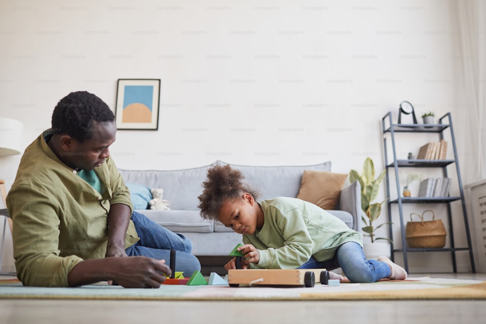 Retrato de vista lateral de un hombre afroamericano jugando con su hija mientras está sentado en el suelo en el acogedor interior de la casa, espacio de copia