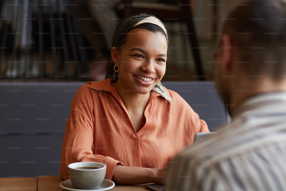 Portrait of young African-American woman smiling and looking at partner while enjoying business meeting at cafe table, copy space