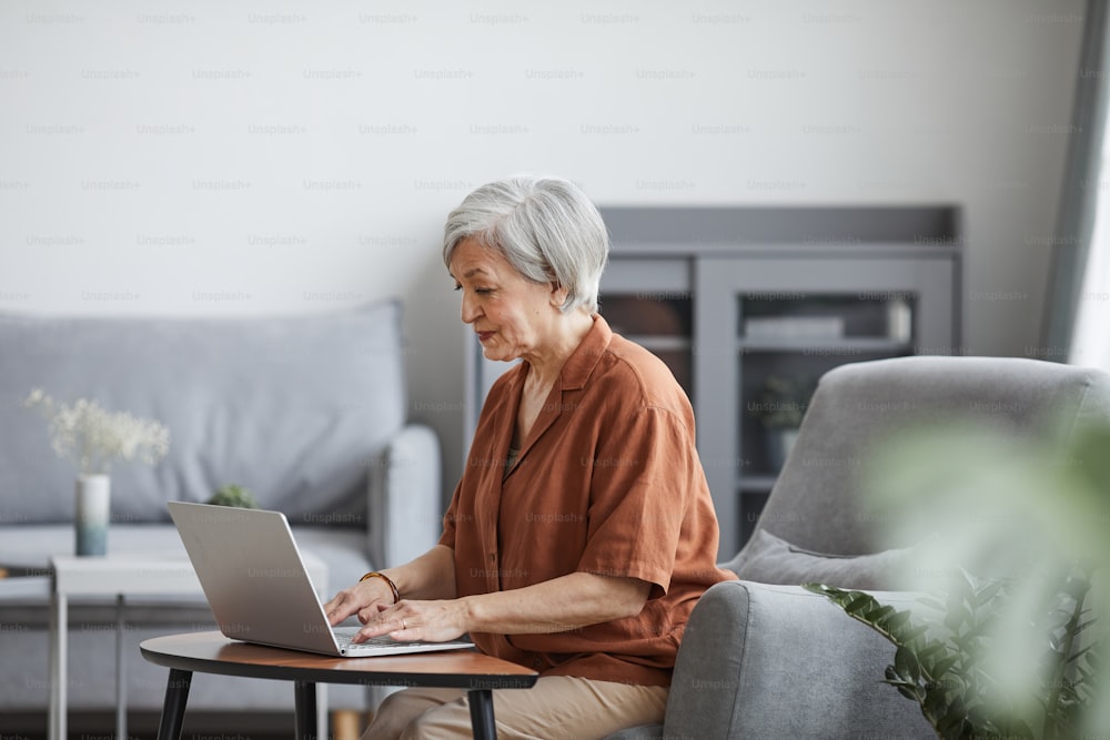 Retrato da vista lateral da empresária sênior que usa o laptop enquanto trabalha em casa no interior mínimo, espaço de cópia