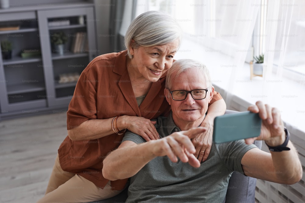 Hochwinkel-Porträt eines glücklichen älteren Paares, das das Smartphone zu Hause zusammen nutzt und sich umarmt