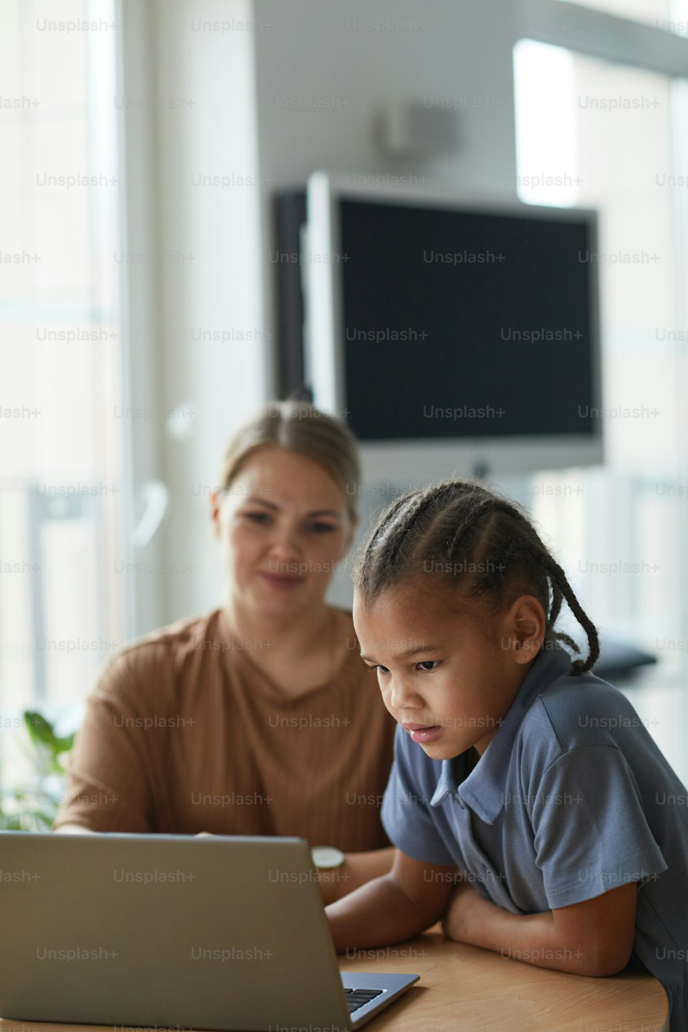 Vertical portrait of cute black girl looking at laptop screen with smiling Caucasian mother in background