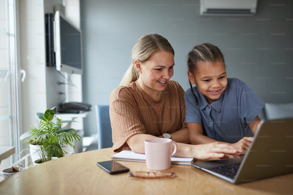 Portrait of multiethnic family, happy mother and daughter looking at laptop screen while studying together at home, copy space