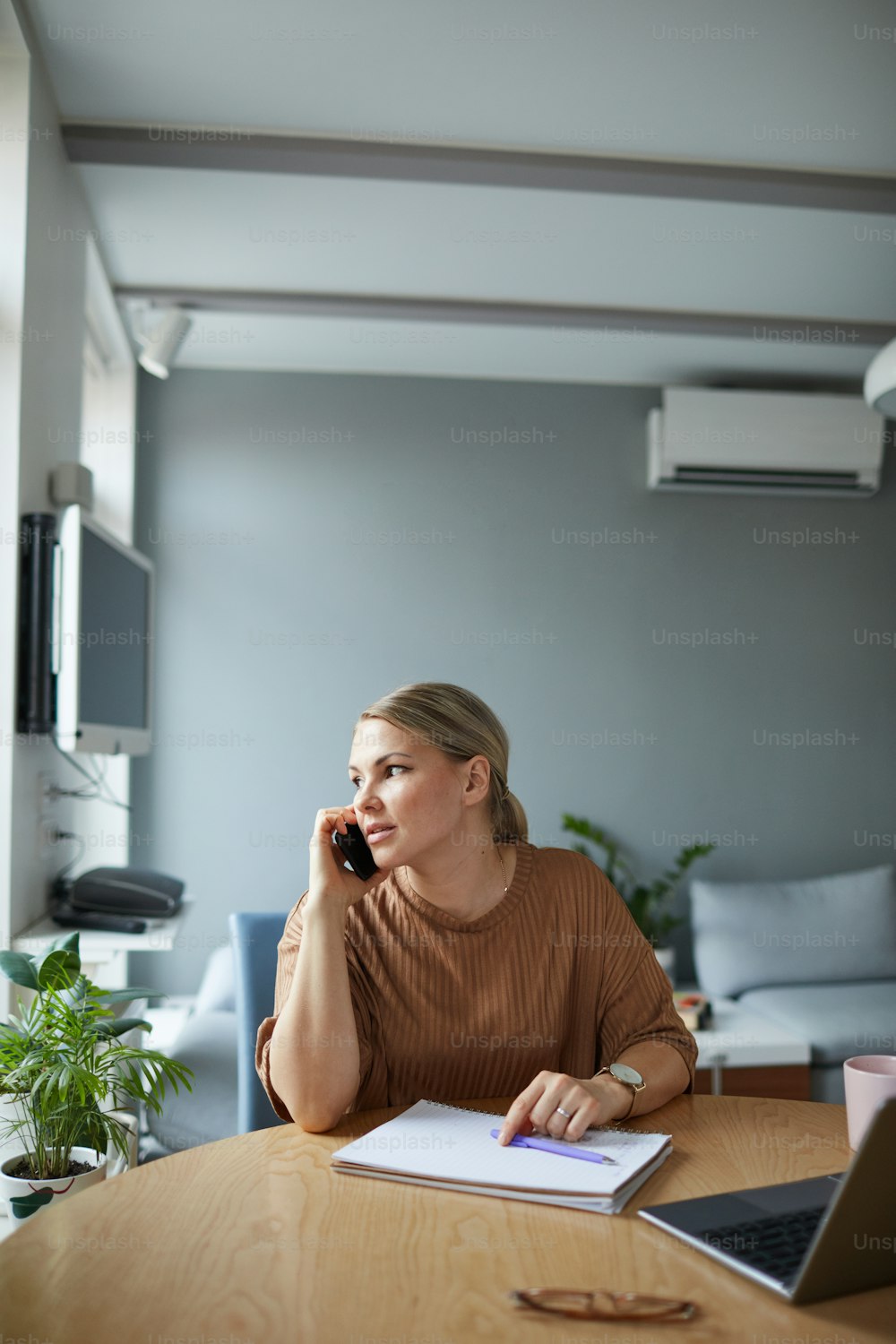 Vertical portrait of young Caucasian woman speaking on phone while working or studying at home in apartment with minimal decor