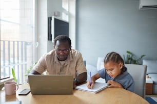 Portrait of young black girl drawing at table with father working from home, copy space