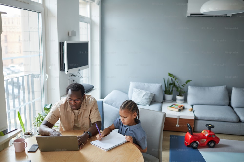 High angle portrait of African American man working from home and using laptop with cute daughter drawing by his side, copy space