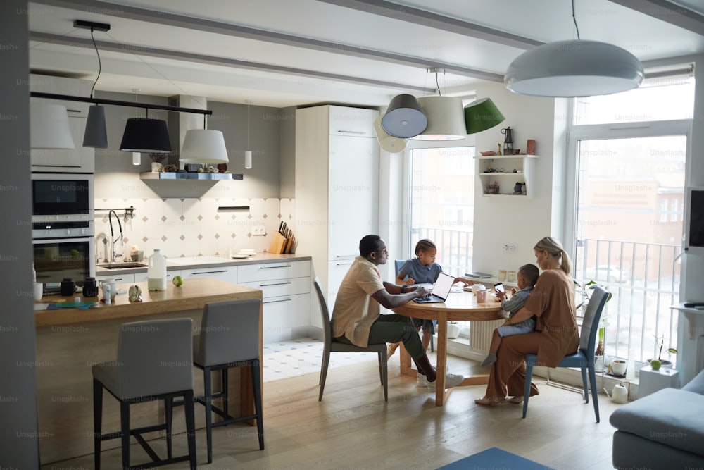 Escena casera de gran angular de la familia multiétnica moderna en la mesa de la cocina, espacio de copia