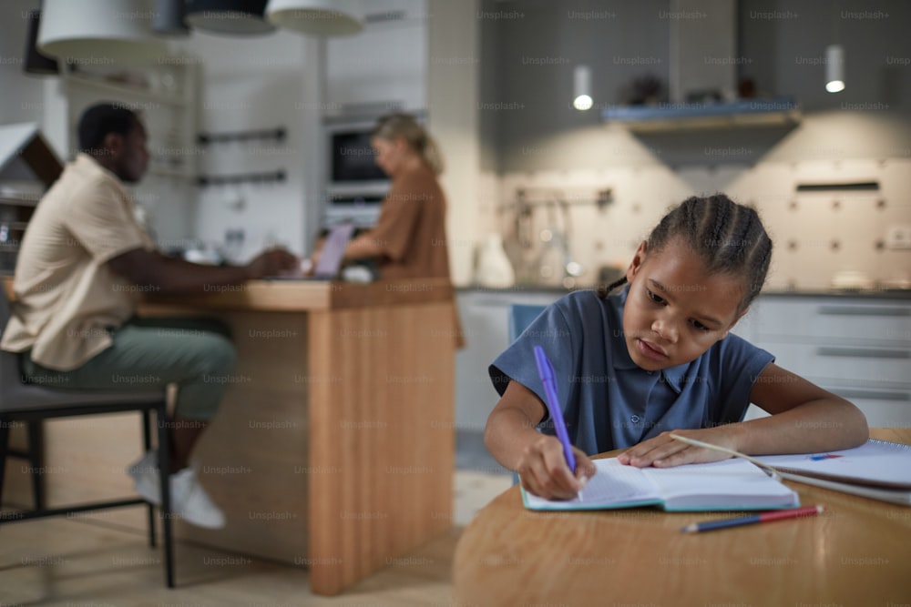Portrait of cute black girl studying at kitchen table with multiethnic parents in background, copy space