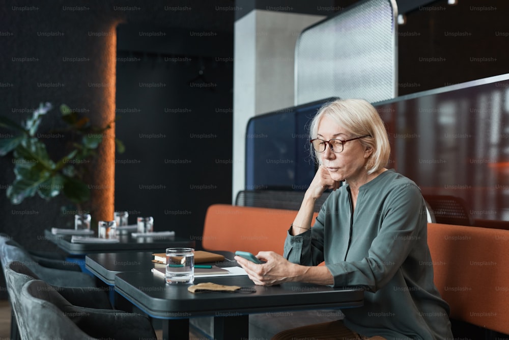 Serious pensive attractive senior lady with blond hair sitting at table in modern cafe in black color and using phone while scrolling on social media