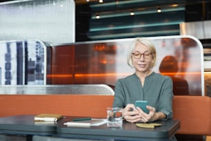 Smiling Caucasian blond-haired senior woman in eyeglasses sitting at table with organizer and checking phone in modern cafe