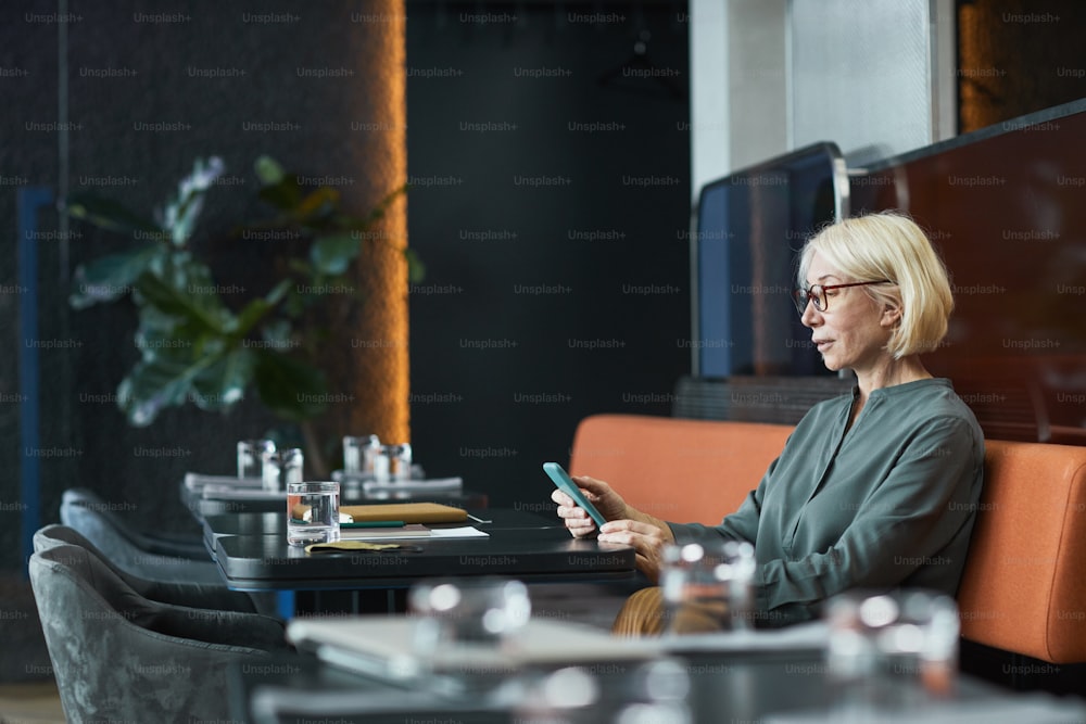 Serious pensive mature lady with blond hair sitting at table in fashionable cafe and checking phone notes
