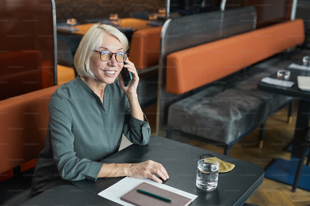 Smiling successful mature businesswoman in eyeglasses and gray shirt sitting at table with organizer and communicating by phone in restaurant