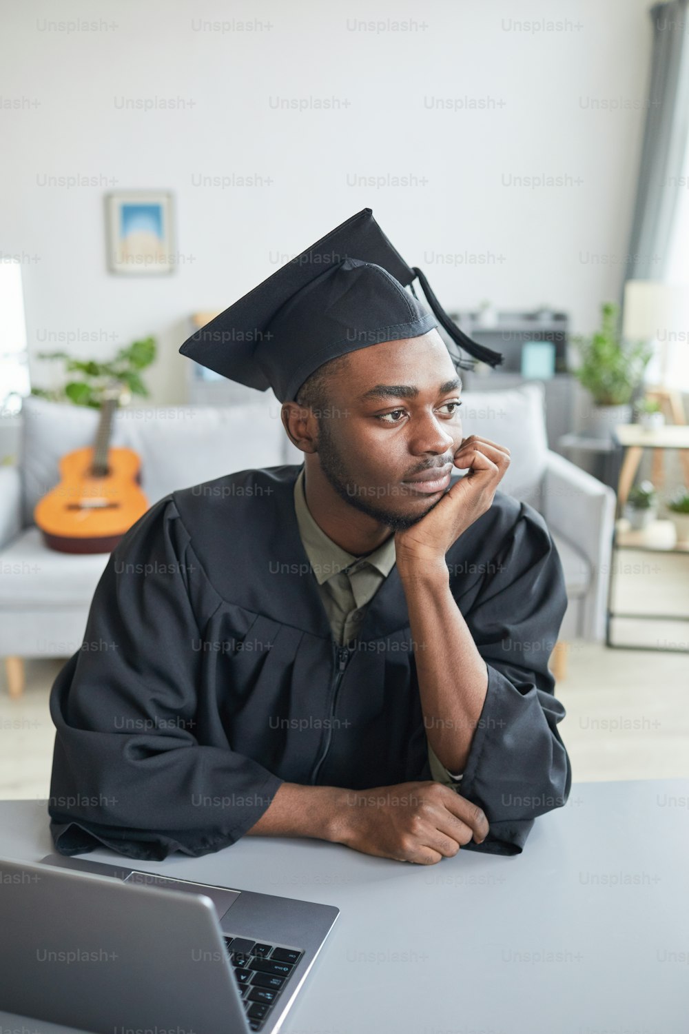 Vertical portrait of pensicve African-American man wearing graduation gown at home