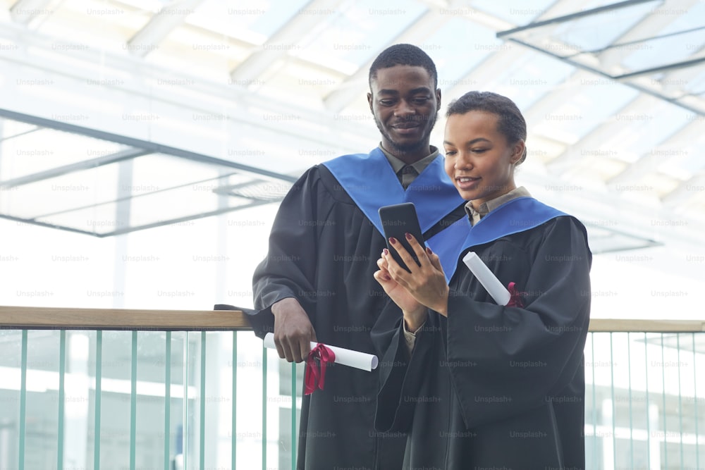 Retrato de cintura para arriba de una joven pareja afroamericana con vestidos de graduación mientras se toma una selfie en el interior de la universidad moderna, espacio de copia