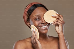 Portrait of carefree African-American woman enjoying skincare and holding natural sponges while looking at camera