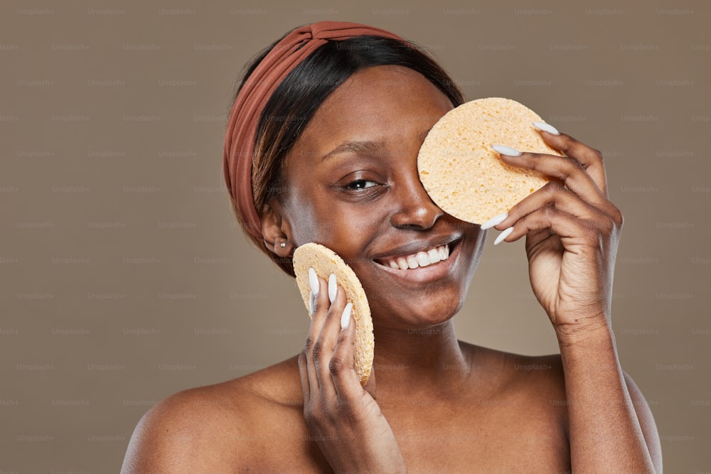 Portrait of carefree African-American woman enjoying skincare and holding natural sponges while looking at camera