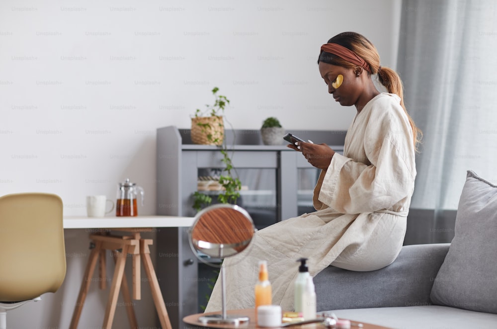 Vue latérale portrait d’une jeune femme afro-américaine appréciant la routine de soins de la peau à la maison et utilisant un smartphone, espace de copie