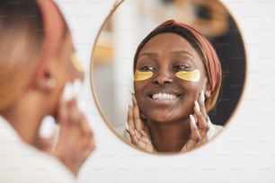 Portrait of beautiful African-American woman enjoying skincare routine at home and looking at mirror, copy space