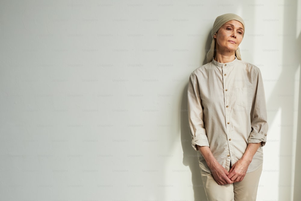 Retrato mínimo de una mujer madura pensativa con pañuelo en la cabeza mientras está de pie junto a la pared blanca a la luz del sol, copia el espacio
