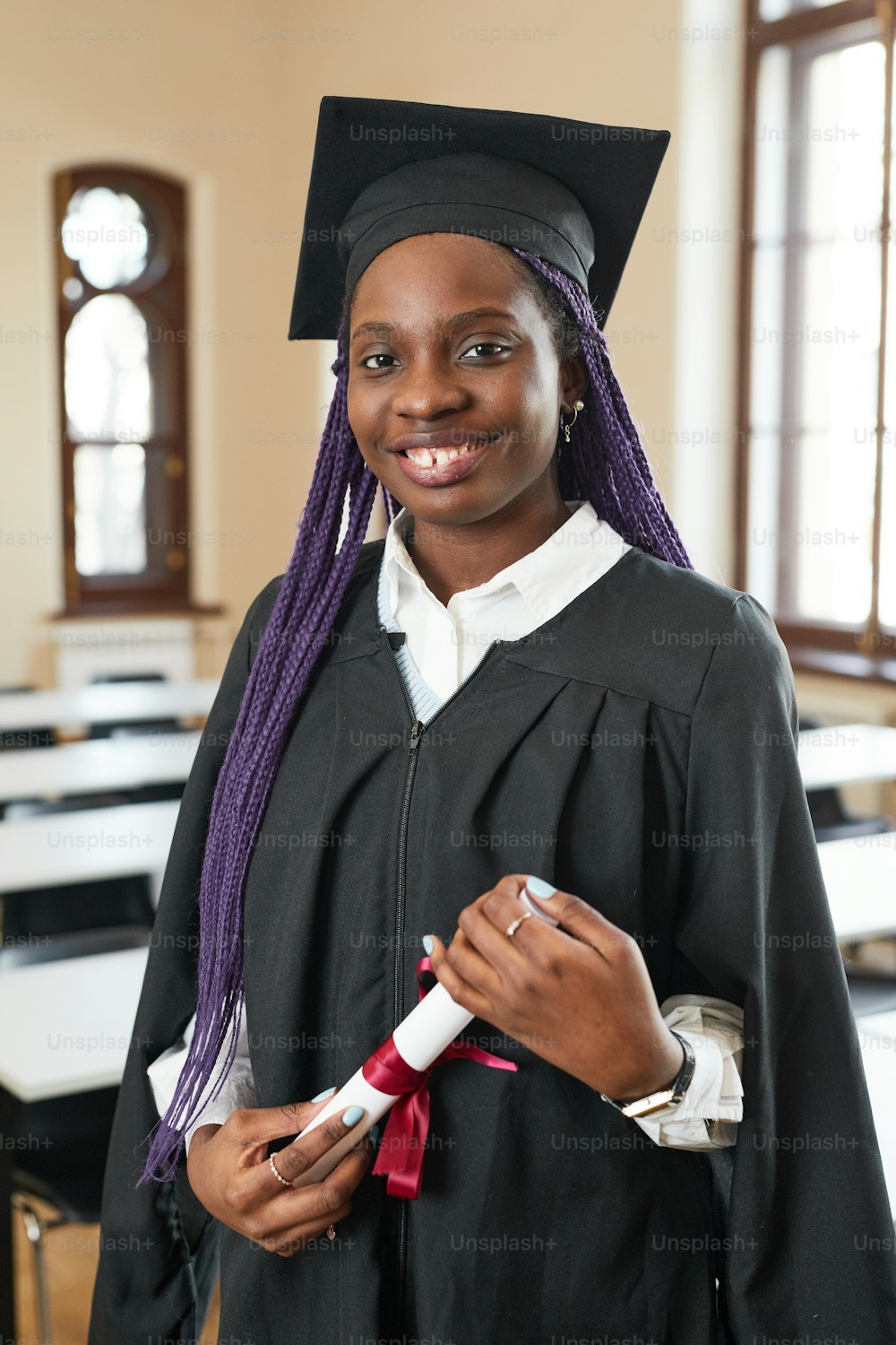 Vertical portrait of African-American young woman wearing graduation robe and smiling at camera while posing in school auditorium
