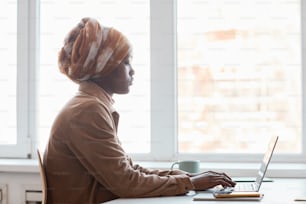 Side view portrait of young African-American woman wearing headscarf while sitting against window in office and using computer, copy space