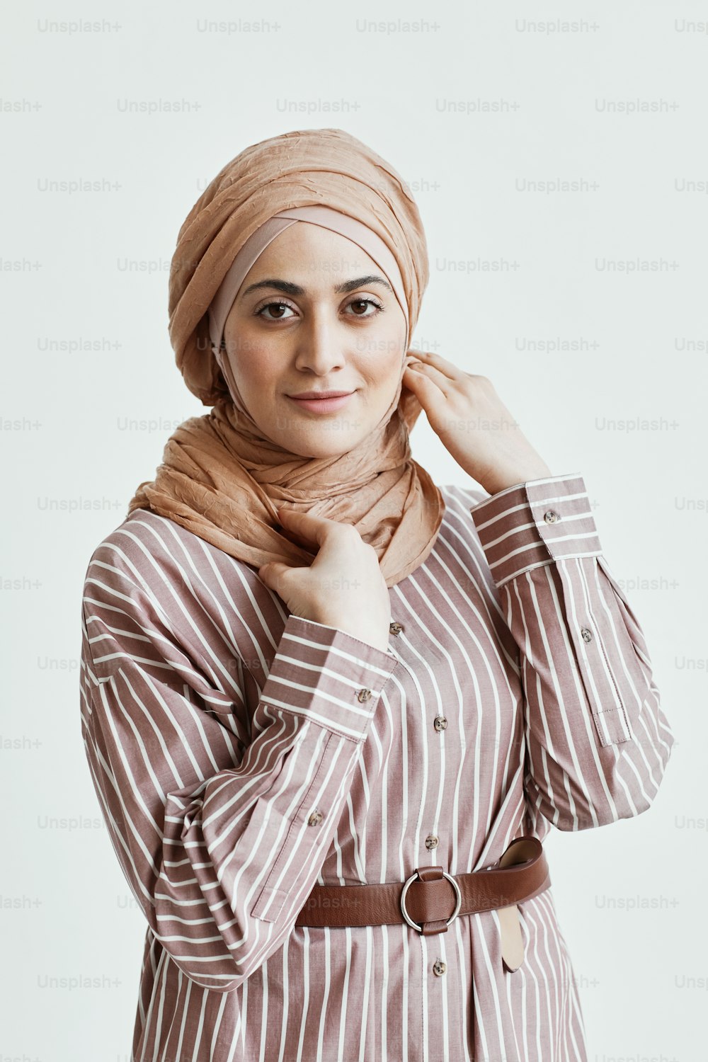 Vertical waist up portrait of modern Middle-Eastern woman looking at camera and wearing headscarf while standing against white wall