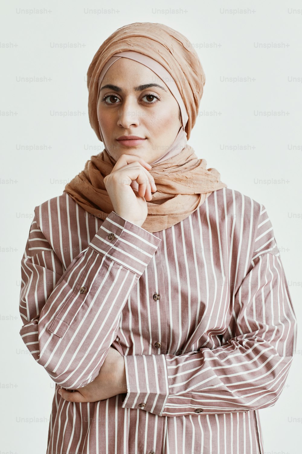 Retrato vertical de cintura para arriba de una hermosa mujer del Medio Oriente mirando a la cámara y usando un pañuelo en la cabeza mientras está de pie contra la pared blanca