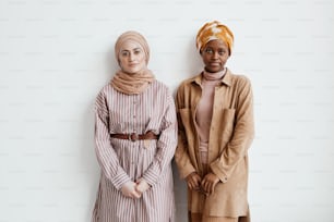 Waist up portrait of two ethnic young women looking at camera while standing against white wall, copy space
