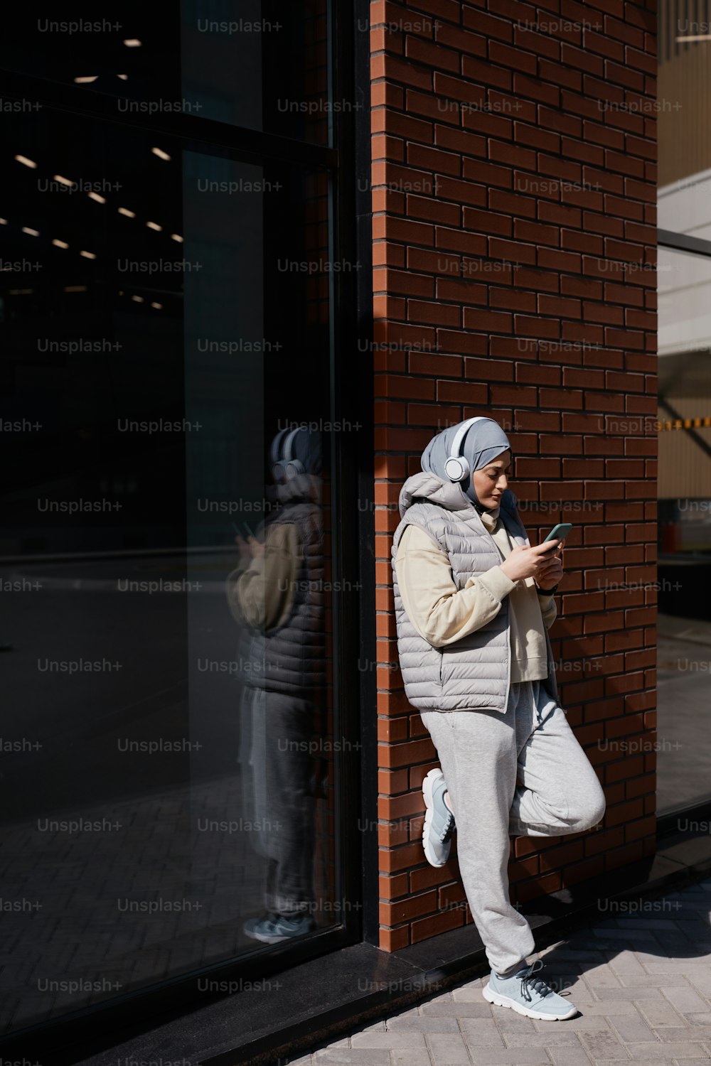 Vertical side view portrait of modern Middle-Eastern woman using smartphone in city lit by sunlight and wearing headphones, copy space