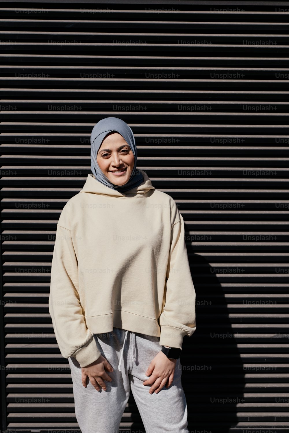 Vertikales grafisches Porträt einer modernen Frau aus dem Nahen Osten, die ein Kopftuch trägt und in eine Kamera schaut, während sie im Sonnenlicht an einer schwarzen Wand steht