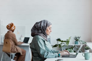 Ritratto di vista laterale della donna moderna del Medio Oriente che indossa il velo in ufficio e utilizza il computer portatile, spazio di copia
