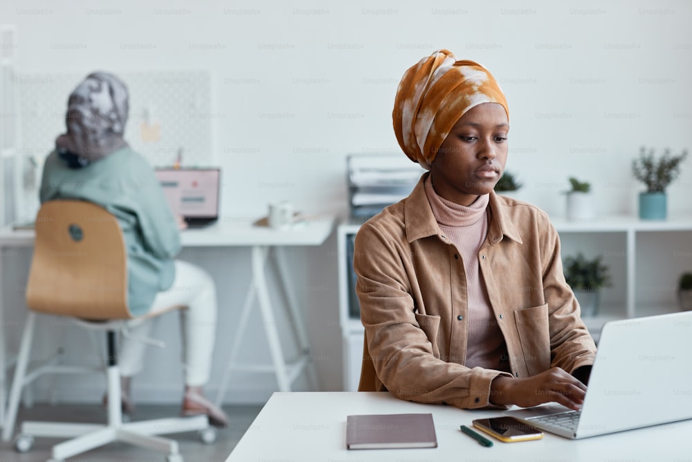 Retrato de una mujer afroamericana moderna con pañuelo en la cabeza en la oficina y usando la computadora, espacio de copia