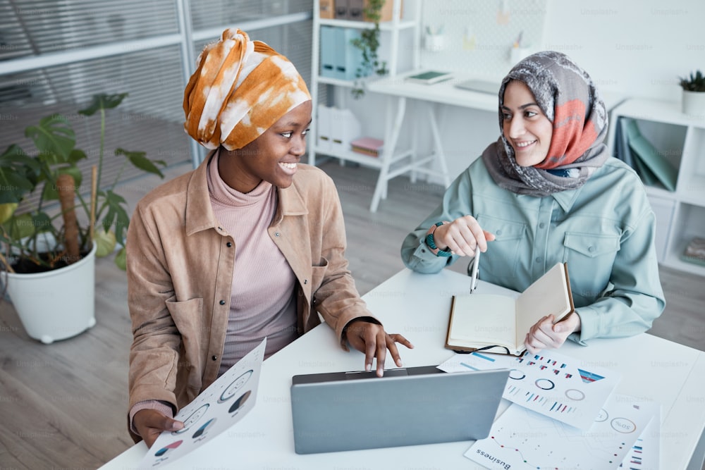 High angle portrait of two ethnic young women wearing headscarf while enjoying work together in office, copy space