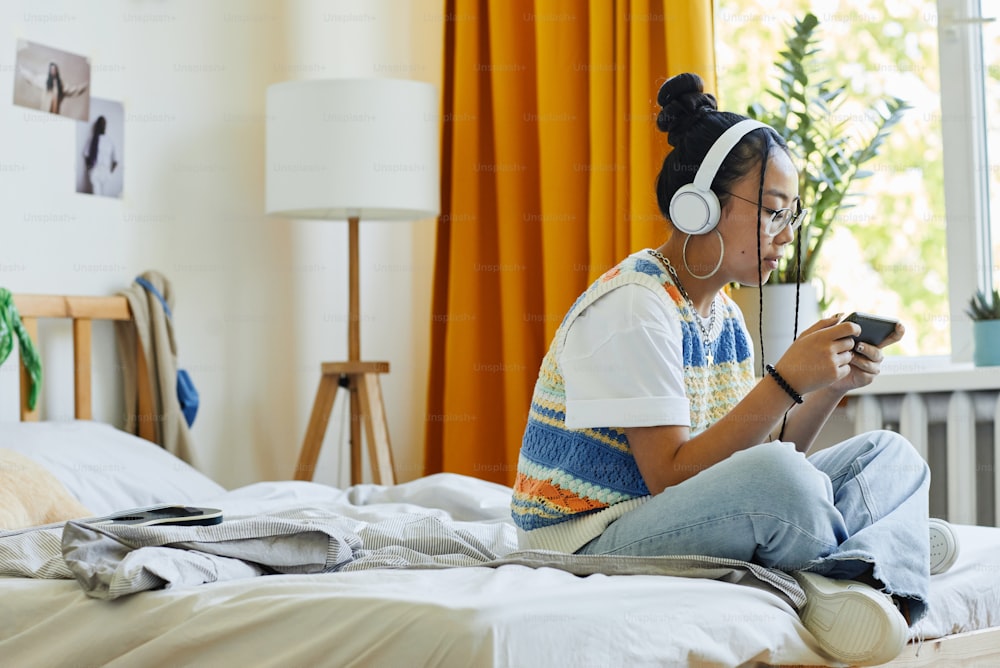 Retrato de vista lateral de una adolescente moderna jugando al teléfono inteligente móvil juego vi mientras está sentada en la cama en una habitación acogedora, espacio de copia