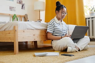Retrato de corpo inteiro de adolescente asiática usando laptop enquanto sentada no chão em casa e estudando, espaço de cópia